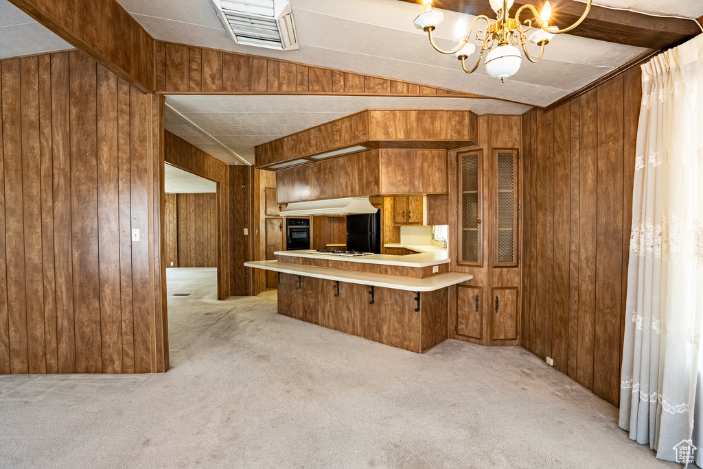 Kitchen featuring light carpet, wooden walls, brown cabinetry, a chandelier, and under cabinet range hood