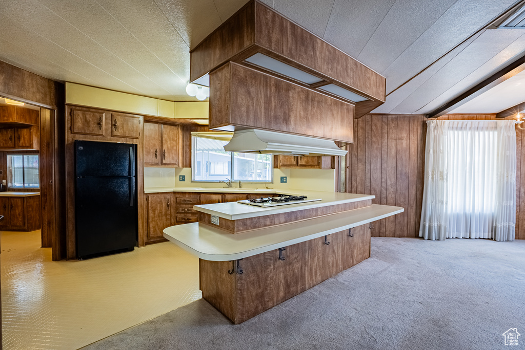 Kitchen featuring freestanding refrigerator, under cabinet range hood, light countertops, wood walls, and a peninsula
