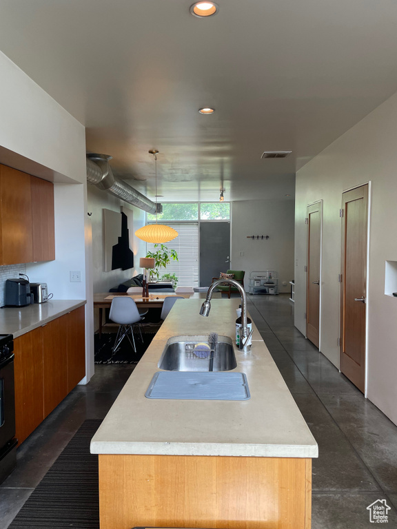 Kitchen featuring a sink, light countertops, recessed lighting, brown cabinetry, and an island with sink