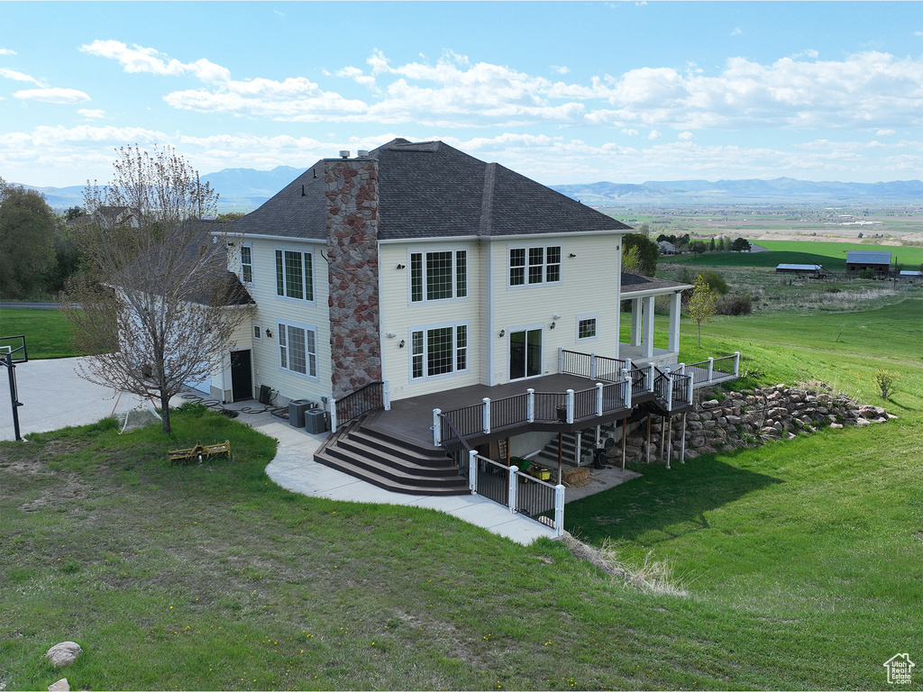 Back of house with a yard, a deck with mountain view, stairs, central air condition unit, and a shingled roof