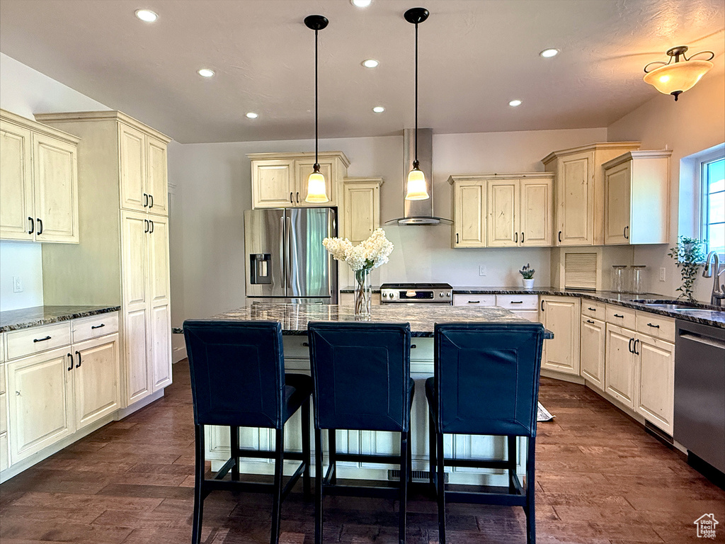 Kitchen featuring stainless steel appliances, cream cabinets, wall chimney range hood, recessed lighting, and a breakfast bar