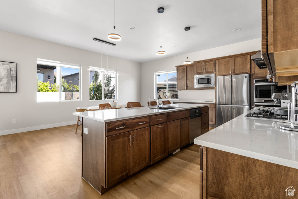 Kitchen featuring appliances with stainless steel finishes, a sink, light wood finished floors, light countertops, and an island with sink