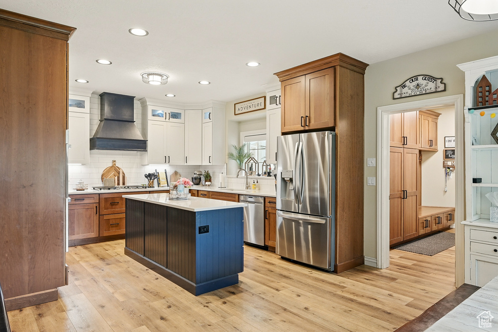 Kitchen with stainless steel appliances, custom range hood, light countertops, backsplash, and light wood finished floors