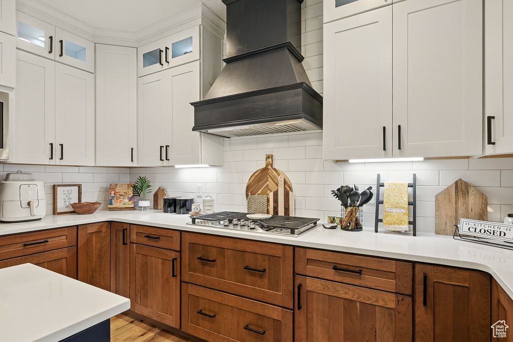 Kitchen with premium range hood, stainless steel gas cooktop, light countertops, glass insert cabinets, and backsplash