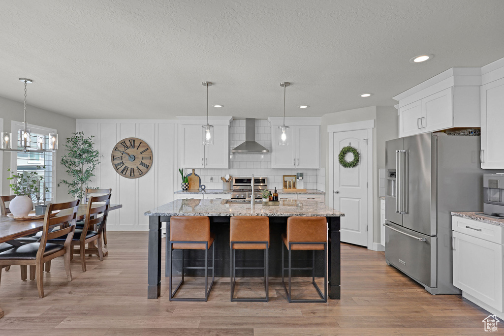 Kitchen featuring wall chimney range hood, stainless steel appliances, a chandelier, a textured ceiling, and white cabinets
