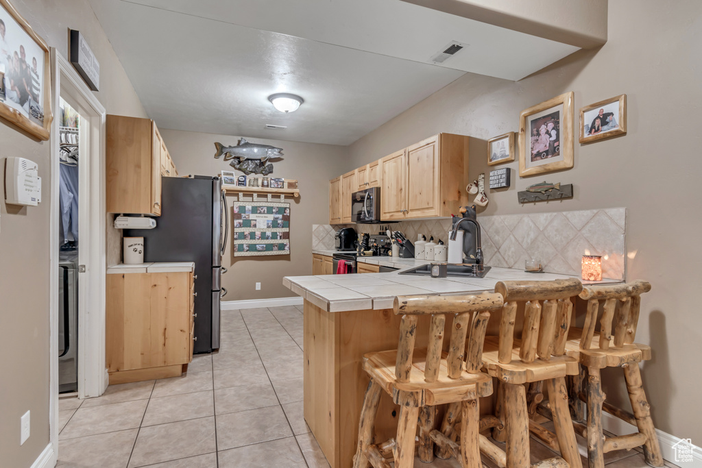 Kitchen featuring stove, tile counters, light brown cabinetry, a peninsula, and decorative backsplash