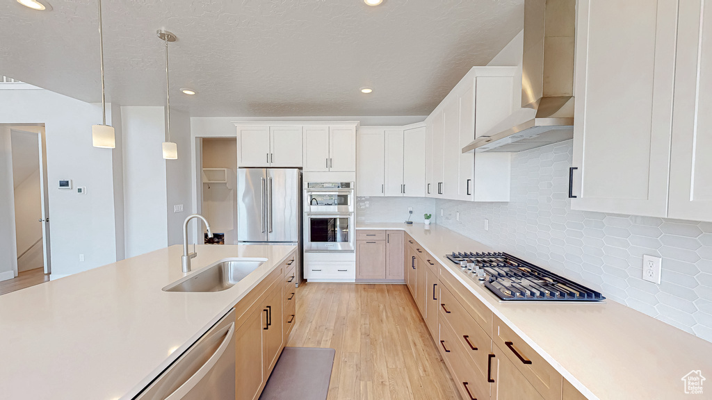 Kitchen with wall chimney range hood, a sink, appliances with stainless steel finishes, light countertops, and light wood-style flooring