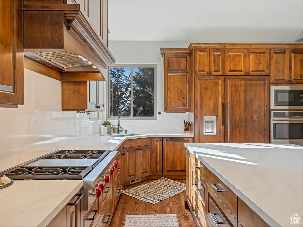 Kitchen featuring stainless steel appliances, a sink, wood finished floors, custom exhaust hood, and brown cabinetry