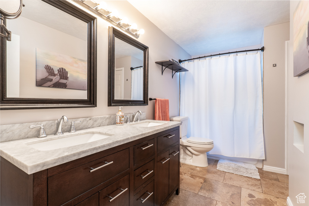 Full bath with double vanity, toilet, baseboards, stone finish flooring, and a shower with shower curtain