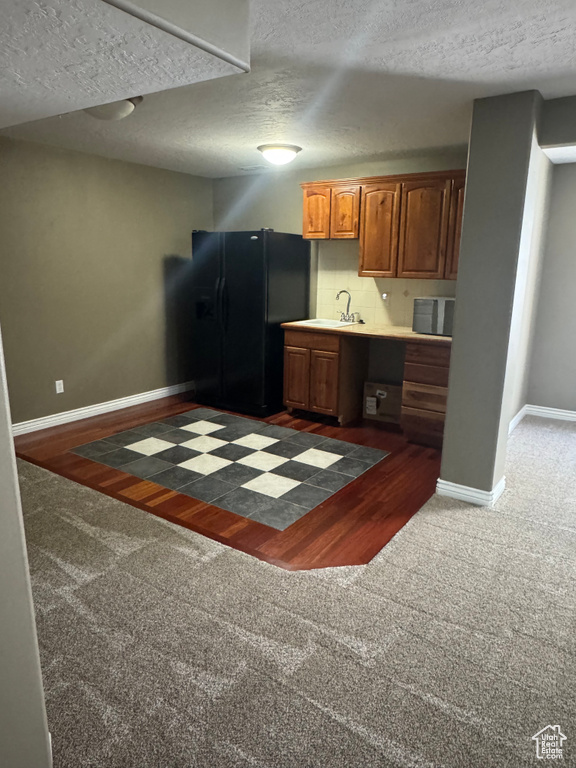 Kitchen featuring dark colored carpet, black fridge, baseboards, brown cabinetry, and a textured ceiling