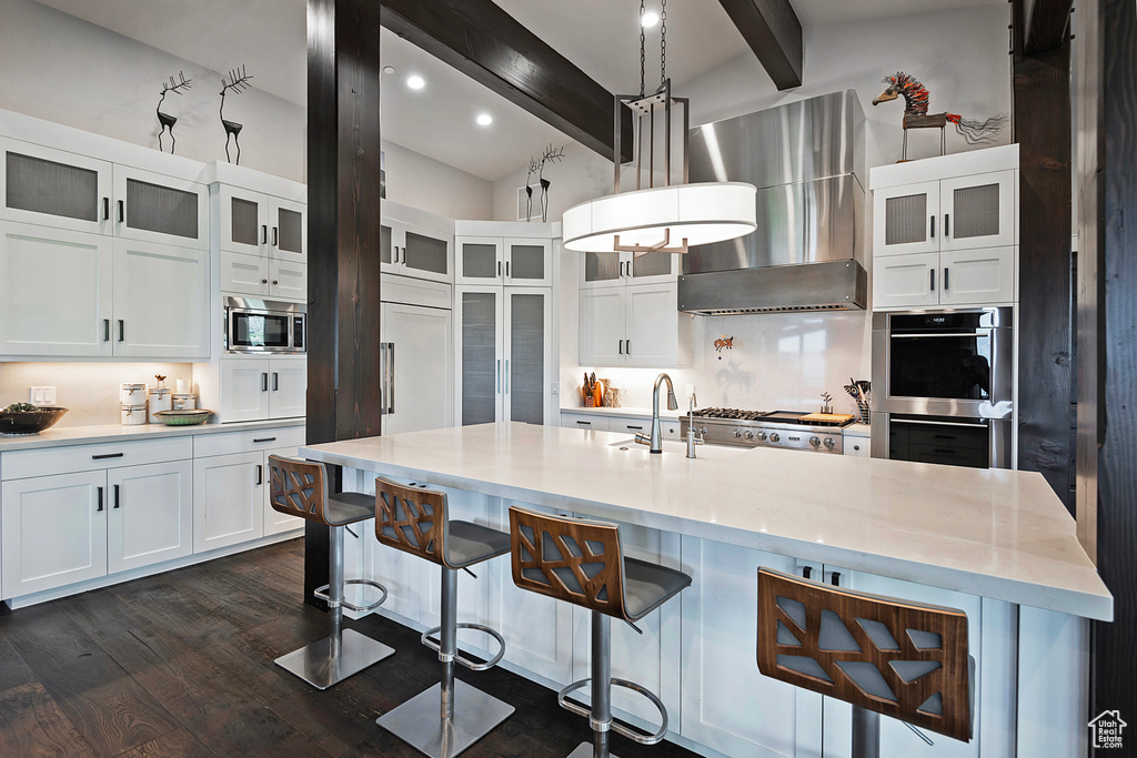 Kitchen featuring wall chimney range hood, a sink, beam ceiling, appliances with stainless steel finishes, and dark wood-type flooring