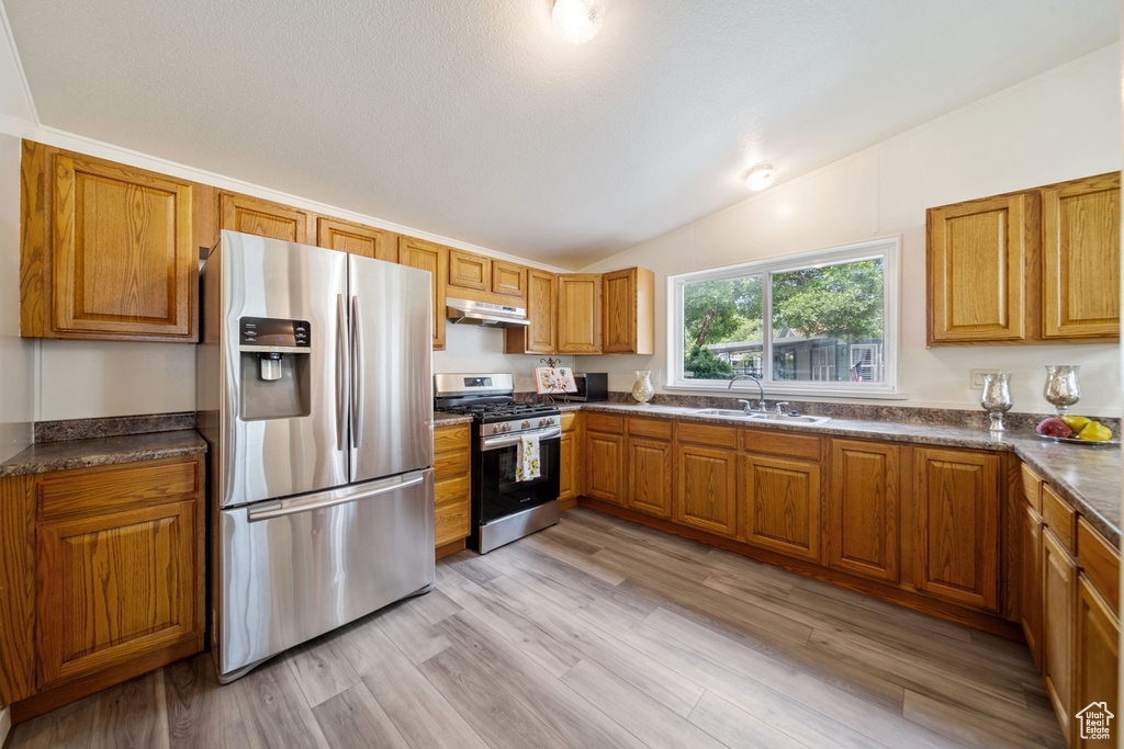 Kitchen featuring appliances with stainless steel finishes, under cabinet range hood, vaulted ceiling, brown cabinets, and a sink