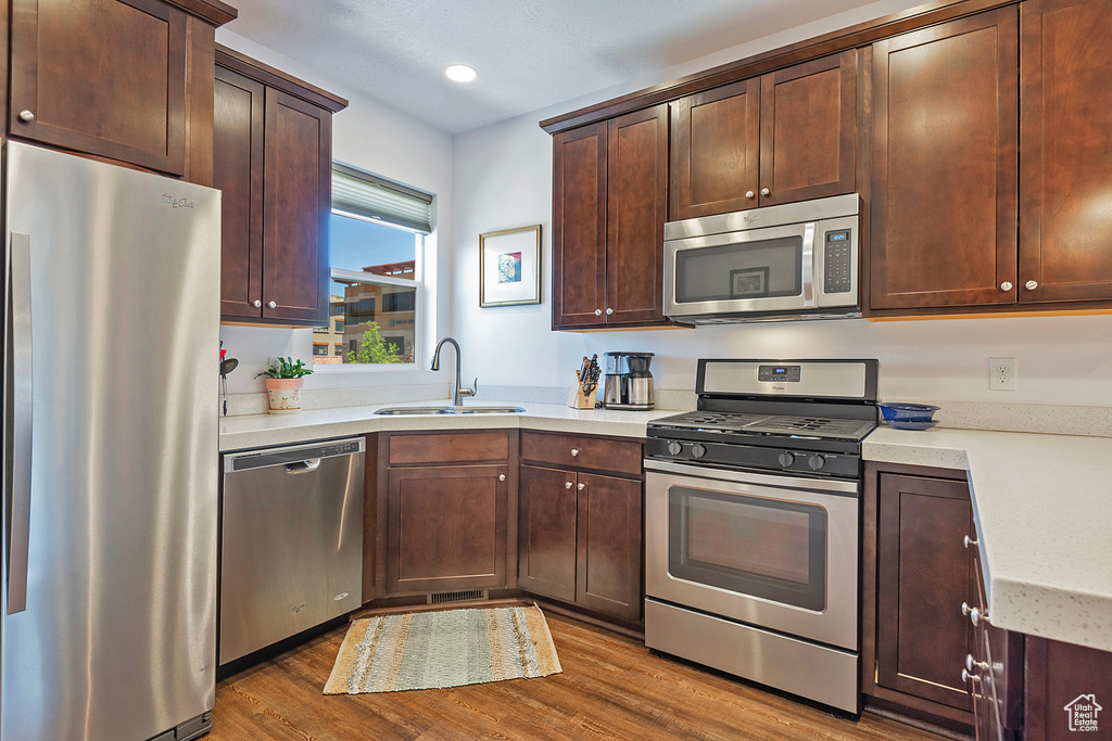 Kitchen with stainless steel appliances, a sink, wood finished floors, light countertops, and dark brown cabinets