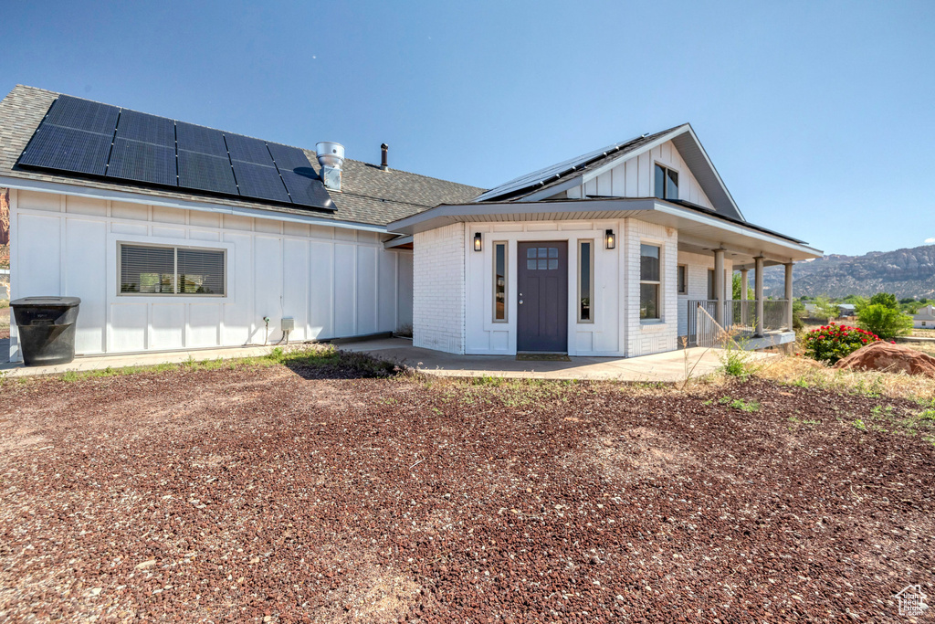 Back of house with board and batten siding, covered porch, roof mounted solar panels, roof with shingles, and brick siding