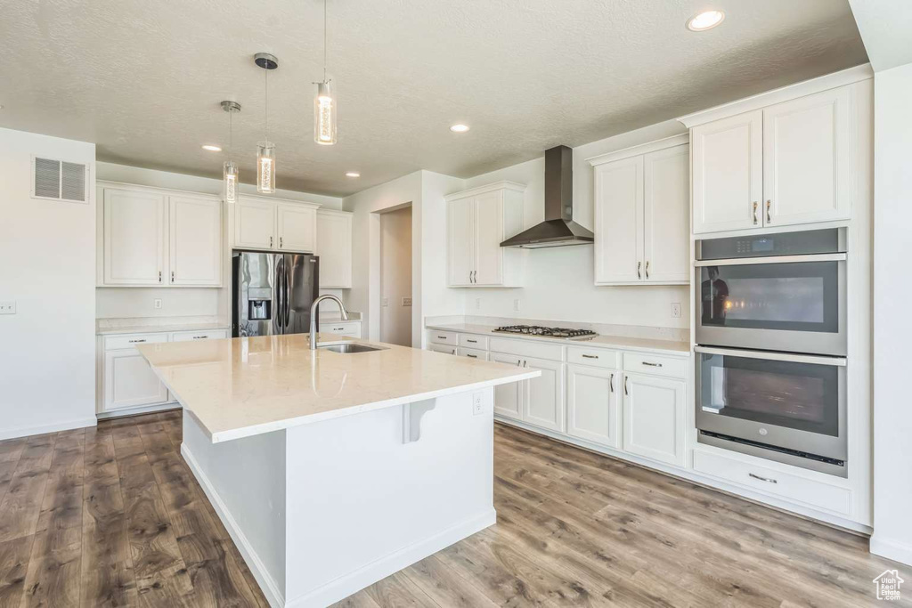 Kitchen with appliances with stainless steel finishes, wall chimney range hood, white cabinets, an island with sink, and dark wood-style floors
