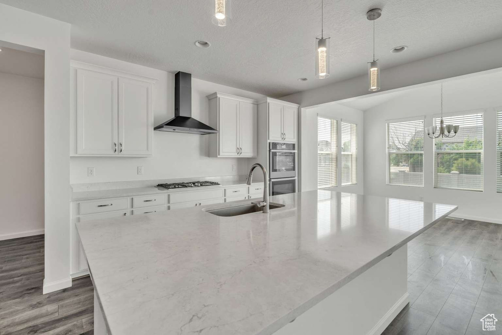 Kitchen featuring wall chimney range hood, stainless steel appliances, an island with sink, white cabinetry, and hanging light fixtures