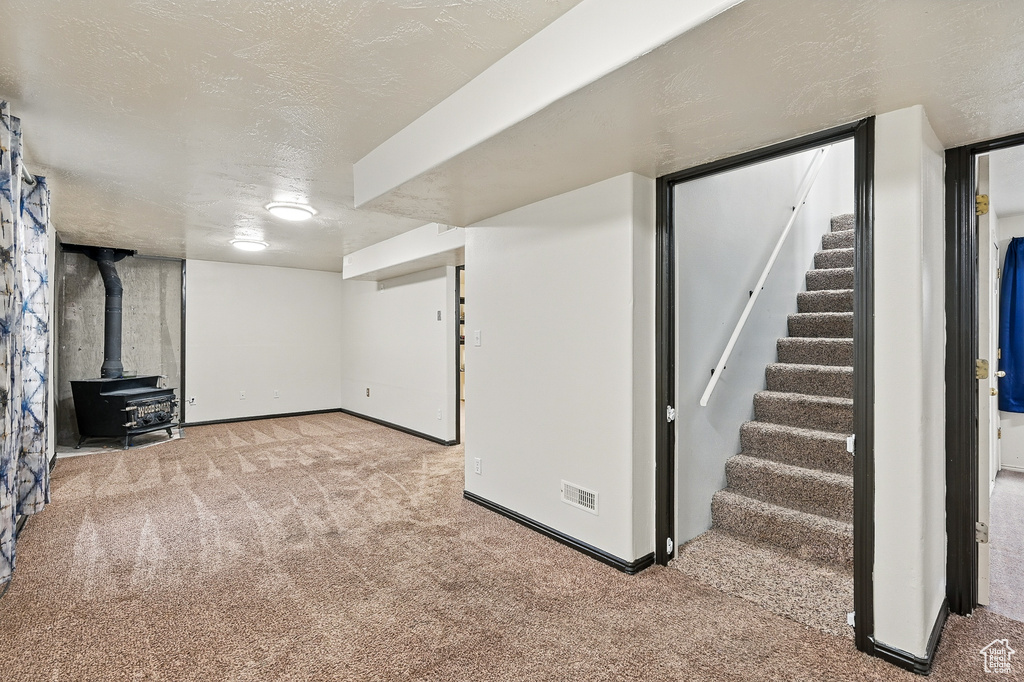 Finished basement featuring carpet flooring, stairway, a wood stove, baseboards, and a textured ceiling
