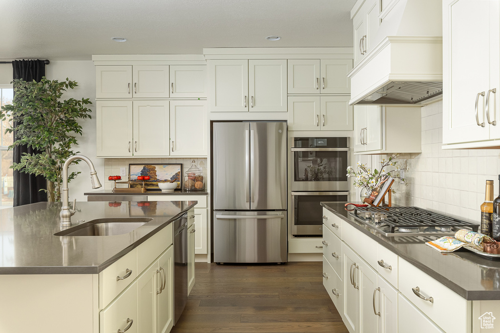 Kitchen featuring appliances with stainless steel finishes, custom range hood, a sink, dark countertops, and tasteful backsplash