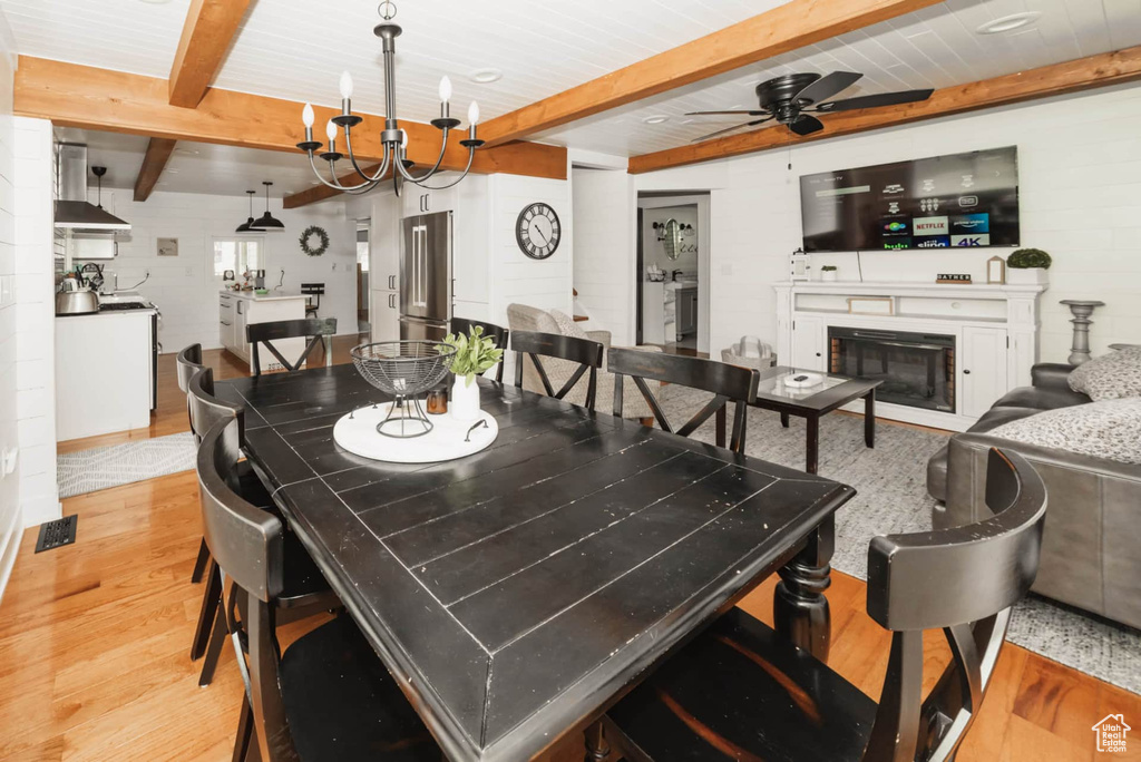 Dining area featuring light wood-type flooring, beamed ceiling, ceiling fan, a glass covered fireplace, and a chandelier