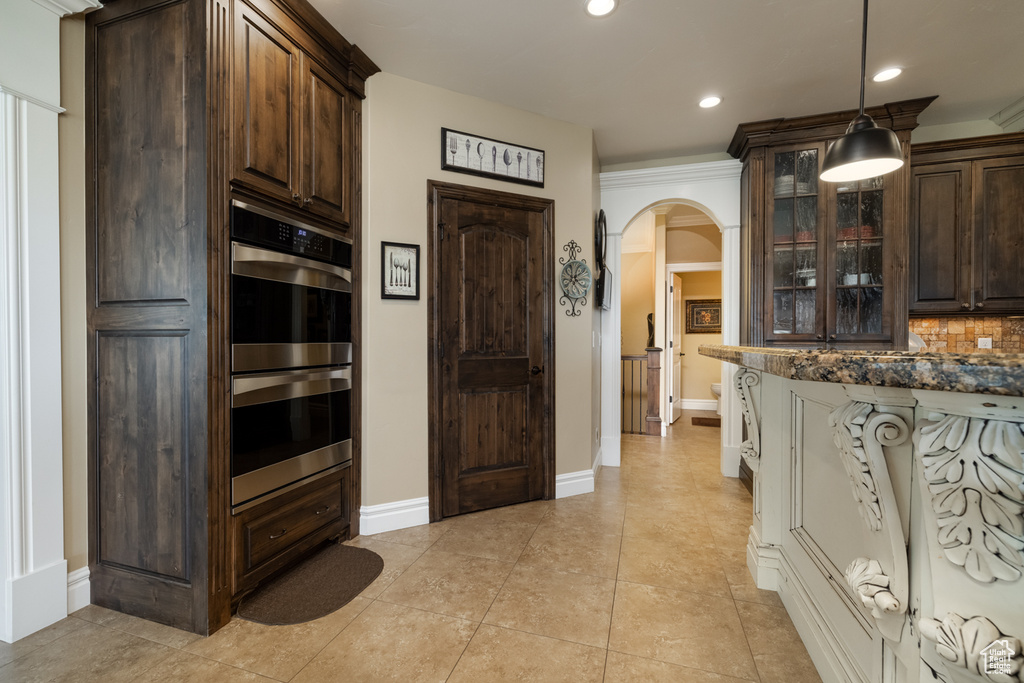 Kitchen featuring double oven, arched walkways, dark brown cabinetry, glass insert cabinets, and backsplash