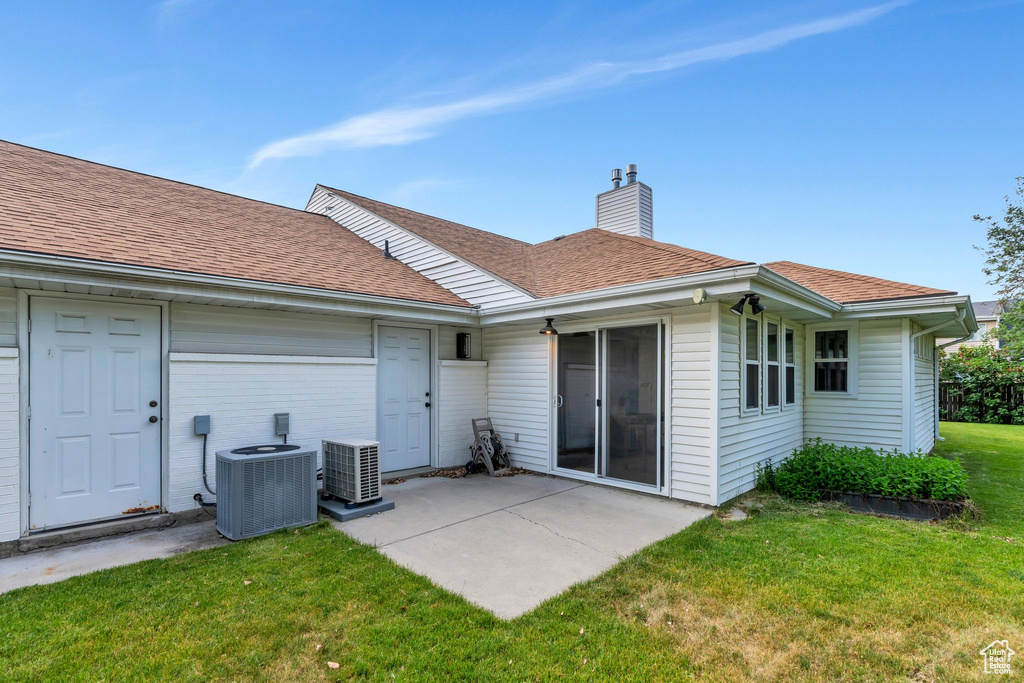 Back of house with cooling unit, a chimney, a yard, a patio area, and a shingled roof
