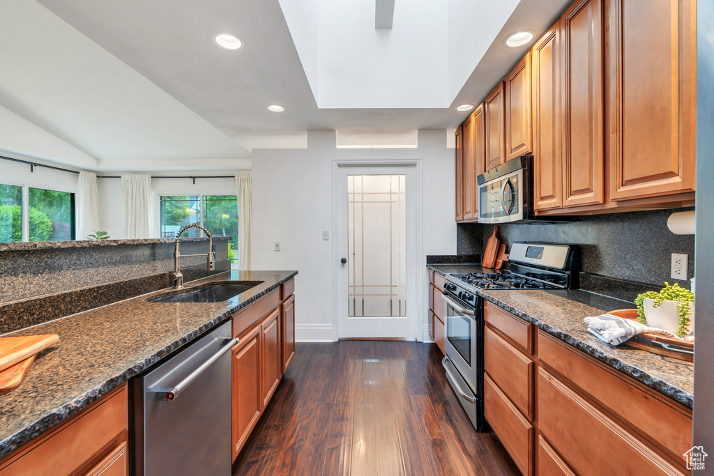 Kitchen featuring stainless steel appliances, a sink, dark wood finished floors, dark stone countertops, and brown cabinetry