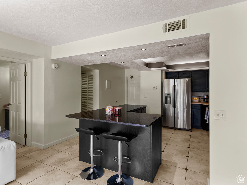 Kitchen featuring stainless steel fridge with ice dispenser, light tile patterned floors, a textured ceiling, a kitchen breakfast bar, and a center island