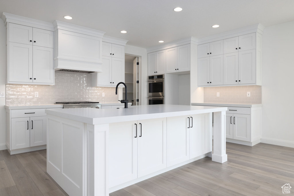 Kitchen with stainless steel double oven, decorative backsplash, a center island with sink, white cabinets, and recessed lighting
