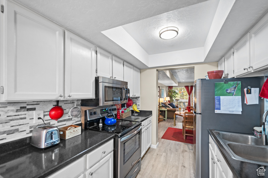 Kitchen with appliances with stainless steel finishes, white cabinetry, light wood-type flooring, tasteful backsplash, and a raised ceiling