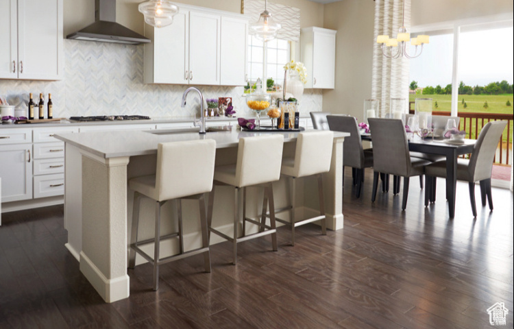 Kitchen with wall chimney exhaust hood, an island with sink, backsplash, and light countertops