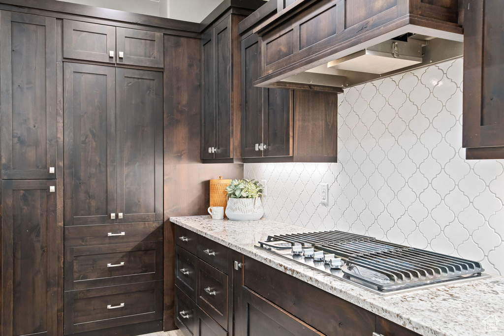 Kitchen featuring stainless steel gas cooktop, dark brown cabinets, decorative backsplash, light stone countertops, and premium range hood