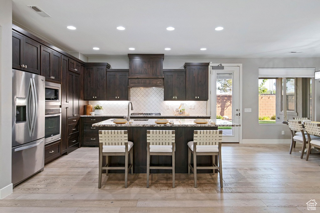 Kitchen featuring appliances with stainless steel finishes, tasteful backsplash, light wood finished floors, dark brown cabinets, and an island with sink