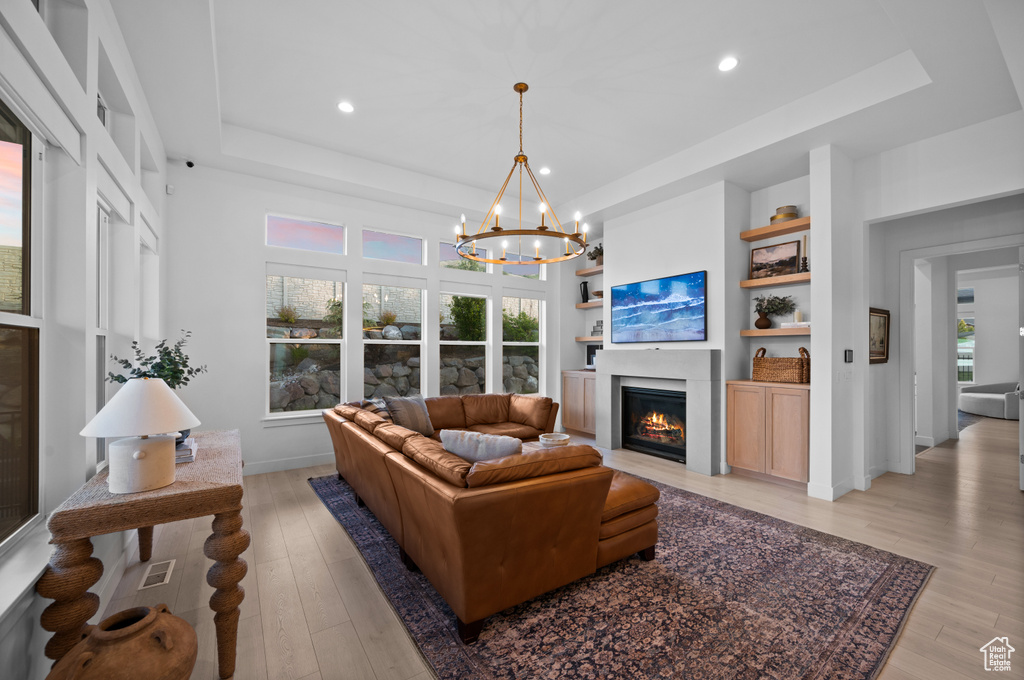 Living room featuring a raised ceiling, light wood-style flooring, a fireplace with flush hearth, a chandelier, and recessed lighting