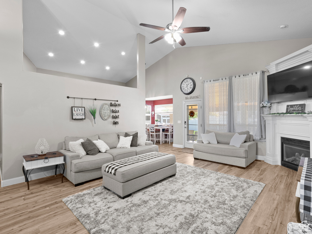 Living room featuring ceiling fan, high vaulted ceiling, light wood-style flooring, a glass covered fireplace, and baseboards