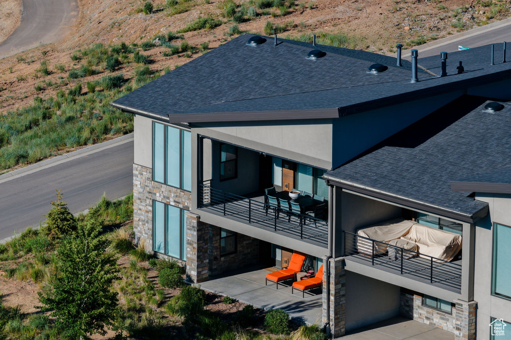 Back of property with stone siding, a balcony, roof with shingles, stucco siding, and a patio area