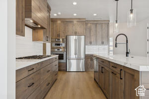 Kitchen with stainless steel appliances, a kitchen island with sink, backsplash, light wood-style floors, and decorative light fixtures