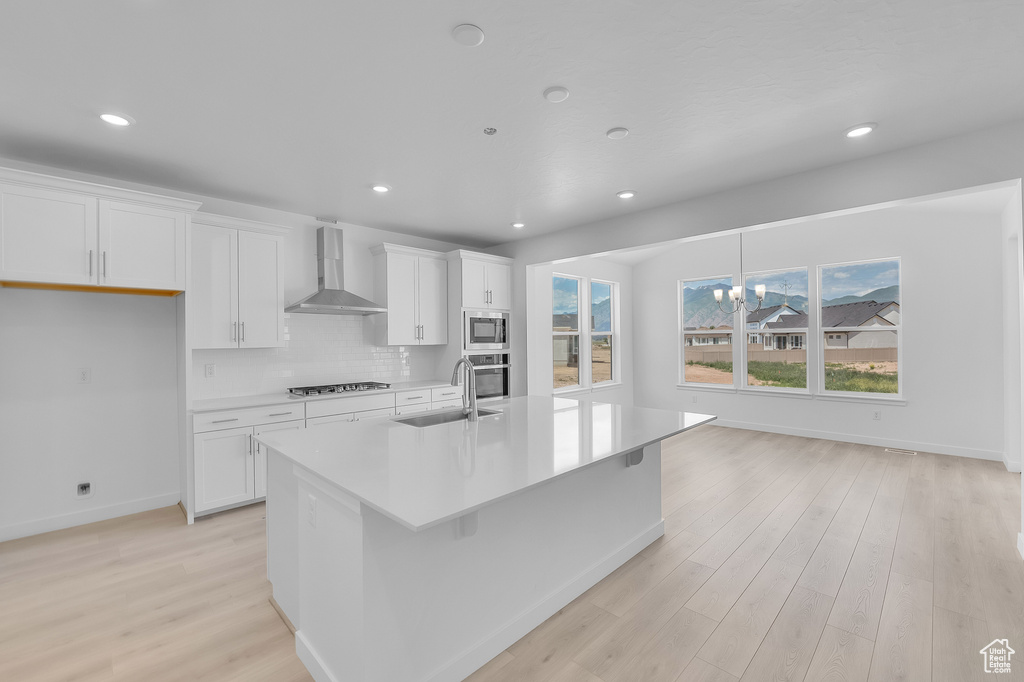 Kitchen featuring wall chimney exhaust hood, appliances with stainless steel finishes, a sink, a kitchen island with sink, and white cabinets