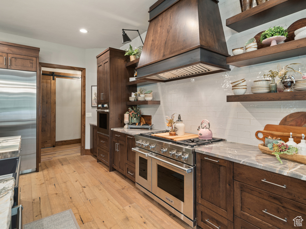 Kitchen with open shelves, stainless steel appliances, custom exhaust hood, backsplash, and a barn door