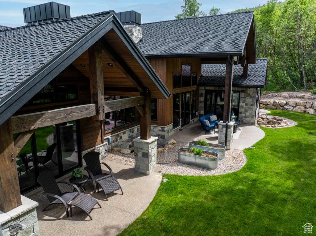 Back of house with a patio area, a chimney, stone siding, a lawn, and roof with shingles