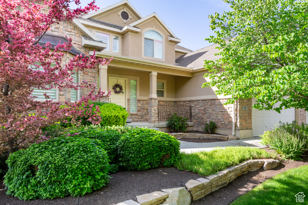 View of front of home with stucco siding, stone siding, a garage, covered porch, and roof with shingles
