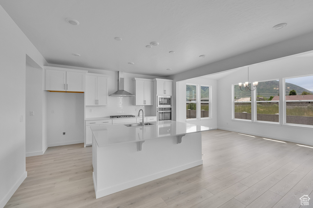 Kitchen featuring wall chimney exhaust hood, a sink, a chandelier, light wood-style floors, and white cabinets