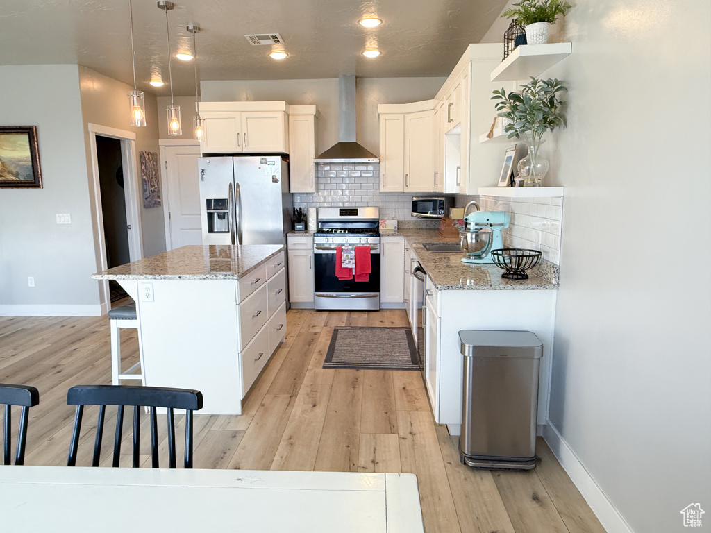 Kitchen featuring stainless steel appliances, wall chimney range hood, a kitchen island, white cabinetry, and light stone counters