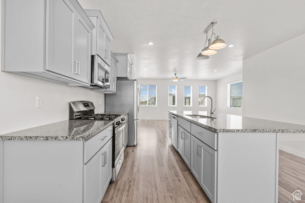 Kitchen featuring stainless steel appliances, a sink, light wood finished floors, ceiling fan, and a center island with sink