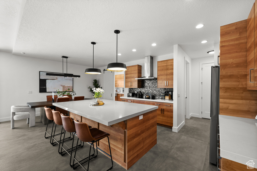 Kitchen with wall chimney exhaust hood, brown cabinets, baseboards, tasteful backsplash, and a spacious island