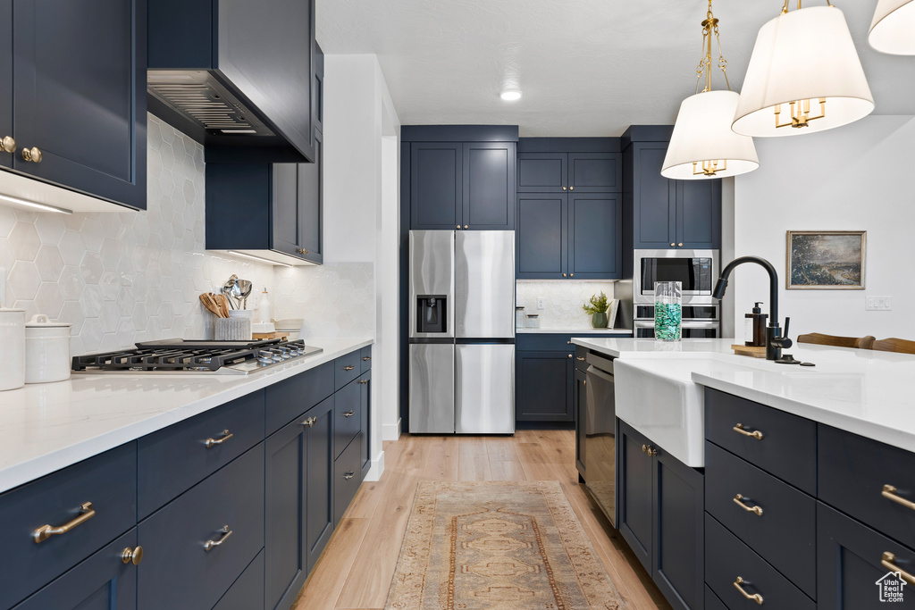 Kitchen with stainless steel appliances, ventilation hood, light wood-style floors, backsplash, and hanging light fixtures