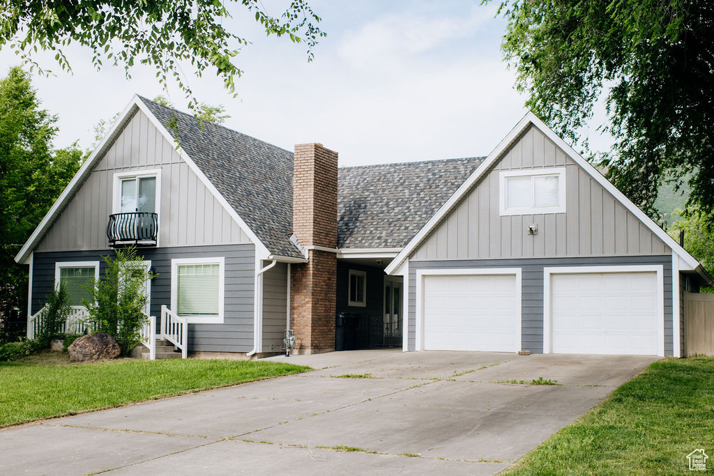 View of front of home featuring a balcony, a chimney, driveway, roof with shingles, and a front lawn