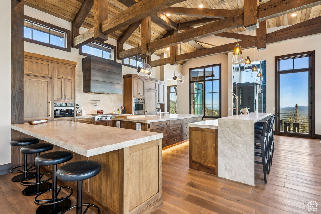 Kitchen featuring appliances with stainless steel finishes, a spacious island, wooden ceiling with exposed beams, a kitchen bar, and wall chimney exhaust hood