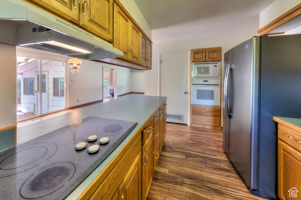 Kitchen featuring white appliances, under cabinet range hood, brown cabinets, dark wood-type flooring, and glass insert cabinets