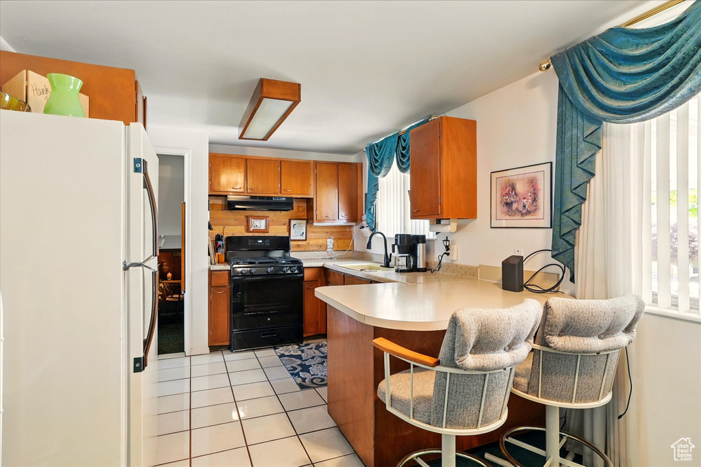 Kitchen featuring freestanding refrigerator, black range with gas cooktop, brown cabinetry, under cabinet range hood, and a peninsula