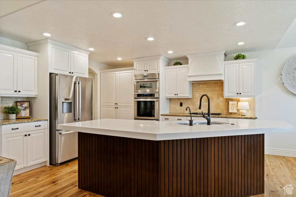 Kitchen with stainless steel appliances, custom range hood, light countertops, a center island with sink, and light wood-style flooring